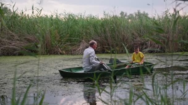 Семейная рыбалка, старший мужчина вместе со своим внуком, плывущим на лодке по озеру между тростником снаружи — стоковое видео