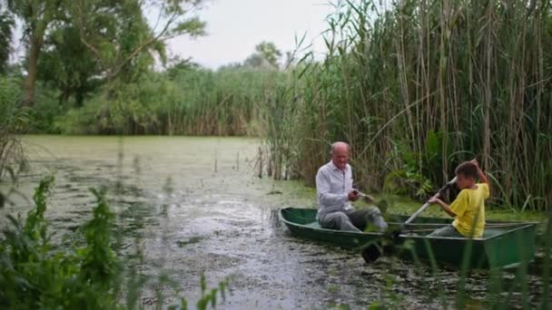 Grandfather and grandson together rowing on river between the reeds on boat, family fishing at summer vacation — Stock Video