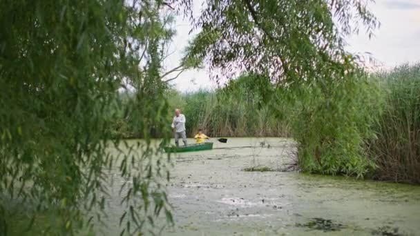Abuelo de pie en barco remando un remo a lo largo del río con su nieto, vista a través de ramas de sauce — Vídeos de Stock