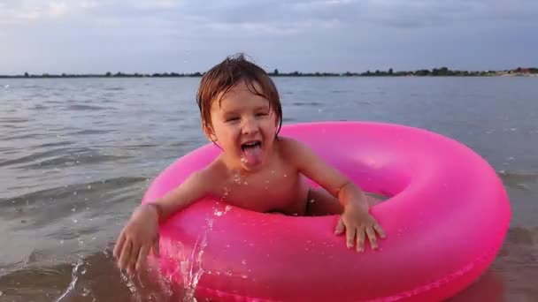 Vacaciones de verano, el niño sentado en el anillo inflable del color rosado está jugando alrededor, niño pequeño que sobresale de su lengua salpica en el mar y las olas saludan de la mano — Vídeos de Stock