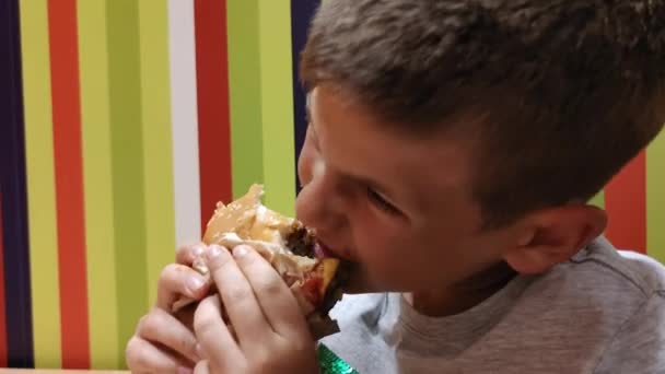 Πεινασμένο μικρό αγόρι απολαμβάνει γρήγορο φαγητό κάθεται στο τραπέζι στο δρόμο καφέ, χαριτωμένο παιδί σβήνει την πείνα με επιβλαβές γεύμα — Αρχείο Βίντεο