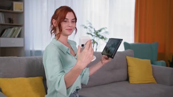 Aplikacja bezpieczeństwa domu, uśmiechnięta właścicielka domu siedzi na kanapie z kamerą i tabletem w rękach — Wideo stockowe