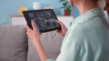 Evdeki güvenlik kamerası sistemini kullanan genç bir kadının arkasını kapatın. Tabletli ve kameralı bir kız koltukta oturuyor.