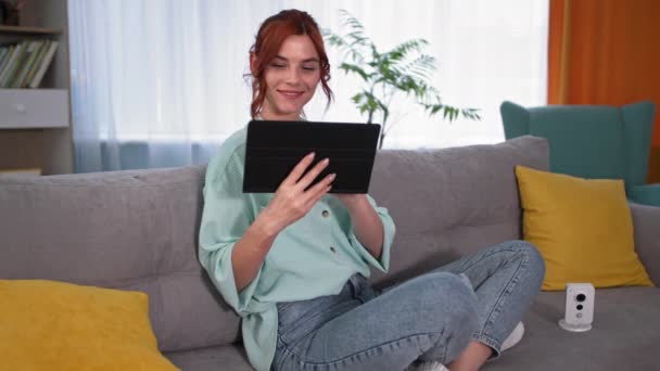 Portraitmädchen überprüft den Video-Feed auf Gadget, junge glückliche Frau studiert die Arbeit der Überwachungskamera mit Tablet-PC, während sie zu Hause auf der Couch sitzt — Stockvideo