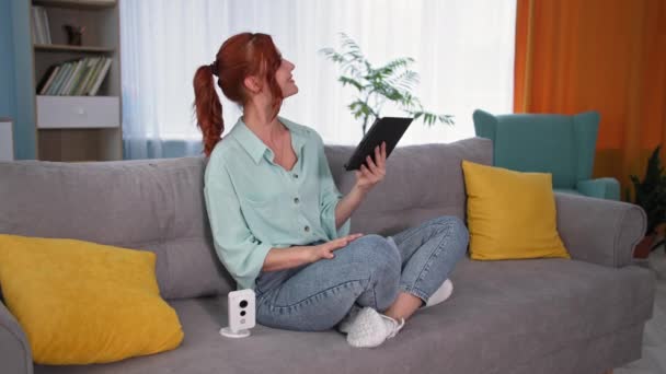 Proprietário controla sistema de vigilância por vídeo com tablet pc, mulher jovem verificando a câmera com gadget em mãos sentadas no sofá dentro de casa — Vídeo de Stock