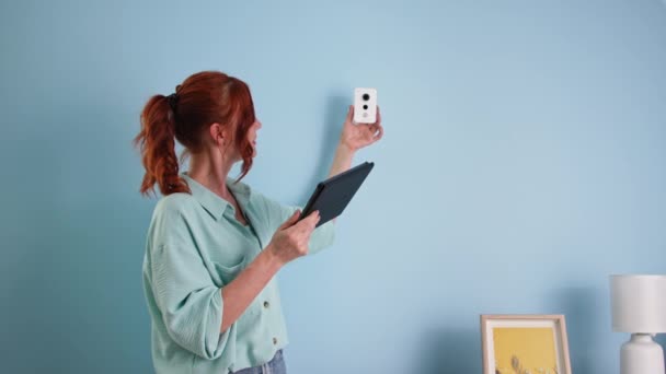 Télécommande à domicile système sur tablette numérique, jeune femme ajuste l'appareil photo à l'aide d'un gadget près du mur et montre un signe similaire — Video