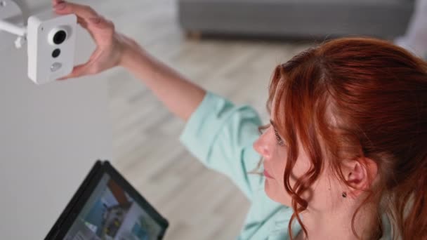 Jeune femme ajustant une caméra de surveillance moderne sur mur blanc à la maison, fille avec tablette dans les mains examine le territoire — Video