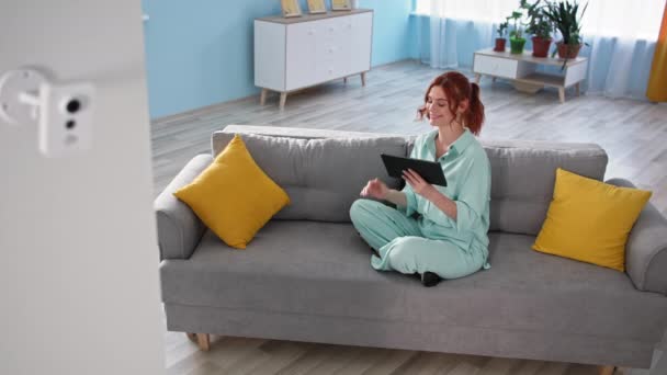 Donna monitoraggio home video sorveglianza su applicazione tablet mentre seduto sul divano, ragazza felice mostrando pollici in appartamento accogliente — Video Stock