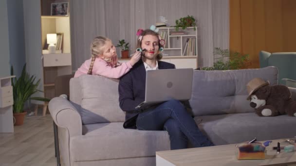 Padre masculino trabaja remotamente en la computadora mientras una niña lo pinta con pinceles de maquillaje y maquillaje durante la pandemia — Vídeo de stock