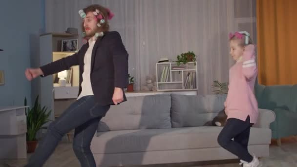 Friendly family, young joyful man and little female child have fun together actively playing and dancing in curlers in living room — Stock Video