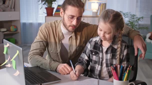 Orang tua dan gadis lucu melakukan pekerjaan rumah bersama-sama selama pelajaran online di sekolah rumah, tersenyum dan melihat kamera — Stok Video