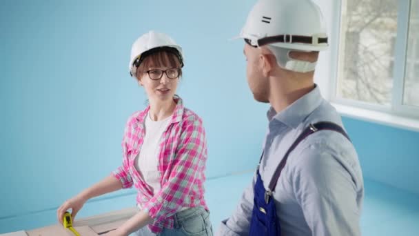 Девушка в строительном шлеме вместе с мужчиной мера ламината будет уложена на пол во время ремонта и установки дома — стоковое видео
