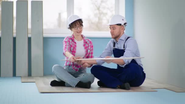 Женщина и мужчина в защитном шлеме обсуждают пол во время ремонта, сидя на пенополистироле — стоковое видео