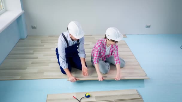 Lavoro di squadra femminile e maschile indossando caschi protettivi che fanno lavori di costruzione e posa pavimenti in laminato durante la ristrutturazione in appartamento — Video Stock