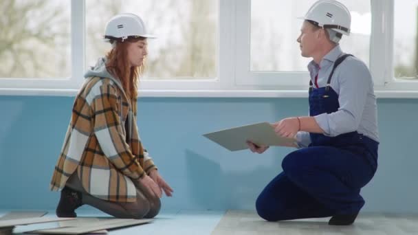 Mężczyzna i kobieta w kaskach budowlanych w procesie układania laminatu podczas remontu podłogi w mieszkaniu — Wideo stockowe