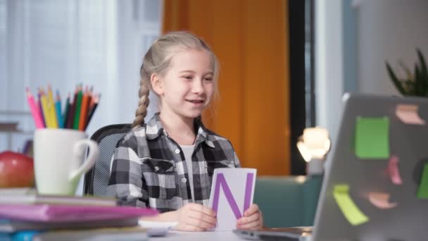 Schoolmeisje is op afstand bezig met de leraar met behulp van moderne technologie en video communicatie op de laptop en toont kaarten met letters op het computerscherm tijdens het zitten — Stockvideo