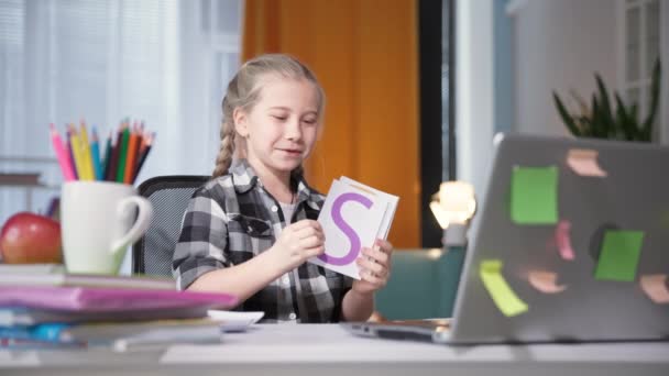 Aluno da escola menina criança faz lição de casa usando webcam no laptop e mostra cartões on-line professor com letras na tela do computador enquanto sentado à mesa em casa — Vídeo de Stock