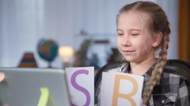 Školačka dělá domácí úkoly s online učitelem a ukazuje karty s písmeny na obrazovce monitoru, zatímco sedí doma u stolu během coronavirus — Stock video