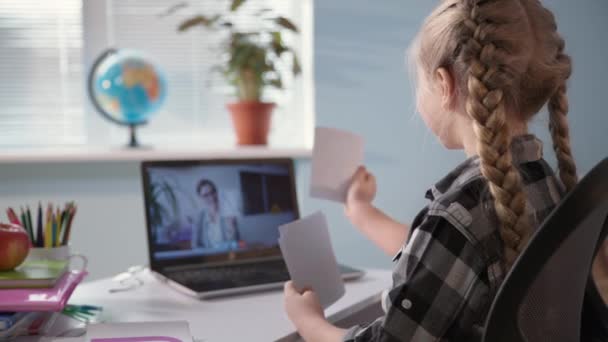 可愛い女子小学生が先生とオンラインでビデオリンクで宿題をしたり、ノートパソコンの画面に手紙でカードを見せたり — ストック動画