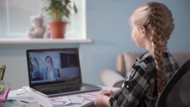 Enfant femelle est engagée dans des cours avec le professeur via vidéo sur ordinateur portable et enseigne les devoirs assis à table dans la chambre — Video