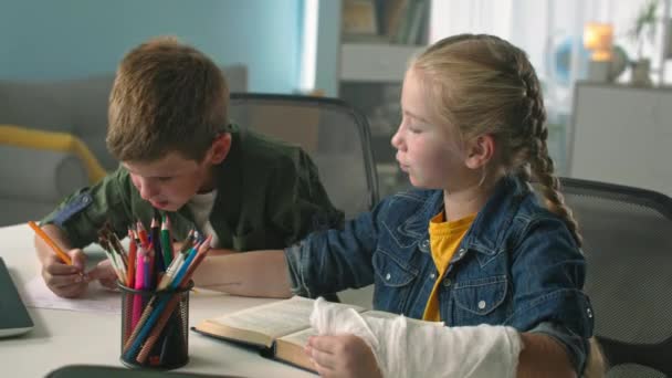 Éducation à la maison, fille avec plâtre jeté sur le bras aide ami avec les devoirs assis à la table avec livre à la chambre — Video