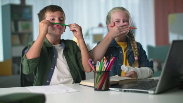 Діти насолоджуються дозвіллям, дівчина з брекетами і кидається на руку обманює хлопчика з другом з кольоровими олівцями, сидячи за столом — стокове відео