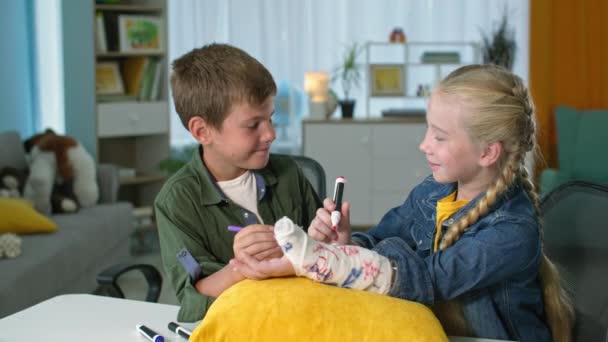 Παιδί φροντίζει και υποστηρίζει τους φίλους, τα παιδιά με πολύχρωμα στυλό τσόχινη άκρη ζωγραφίσει γύψο στο χέρι, αγόρι και κορίτσι δείχνουν αντίχειρες στο σπίτι — Αρχείο Βίντεο