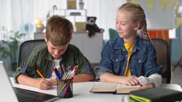 Aprendiendo en casa, niño estudiando para escribir durante la cuarentena con su hermana con yeso en el brazo mientras está sentado en la mesa con el libro — Vídeos de Stock