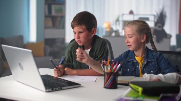 Los niños aprenden en línea, lindo alumno masculino que estudia con una chica con yeso echado en la mano mientras está sentado en casa en la mesa con computadora portátil — Vídeos de Stock