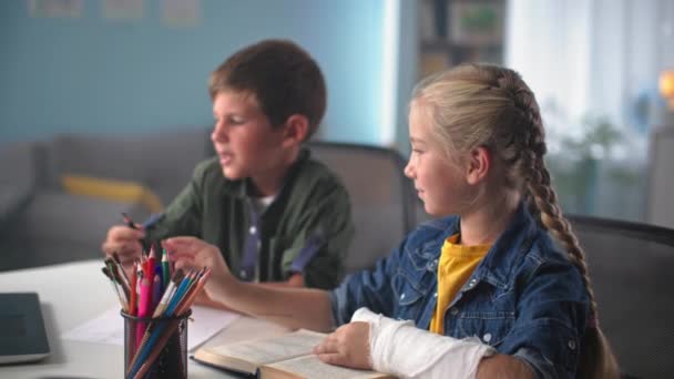 Edukacja domowa, dziewczyna z gipsem na dłoni daje ołówek młodszemu bratu, siedząc przy stole podczas lekcji rysunku — Wideo stockowe