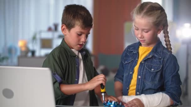 Crianças inteligentes reparar mini quadricóptero com instrumentos perto de laptop, linda menina com um gesso no braço ajuda seu irmão mais novo em casa — Vídeo de Stock