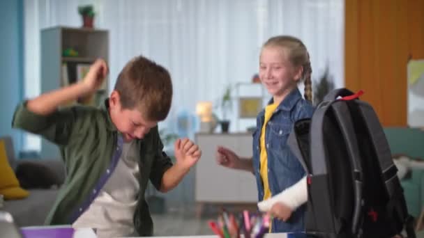 Дети весело танцуют рядом с рюкзаком, девушка с гипсовым гипсом на руке весело проводит время со своим братом дома перед школьными днями — стоковое видео