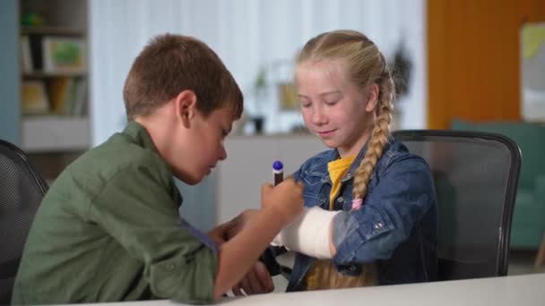 Apoyo y cuidado, niño se basa en elenco de una niña con un brazo fracturado sentado en la mesa en casa — Vídeos de Stock