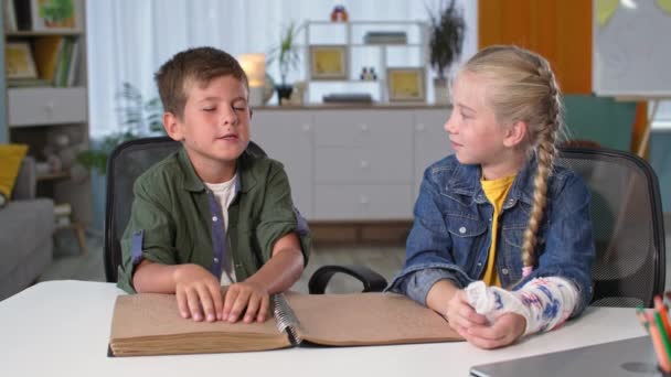 Menino cego está lendo um livro para a menina com um gesso na mão, criança deficiente visual está tocando o braille com as mãos enquanto sentado na mesa ao lado de sua irmã em casa — Vídeo de Stock