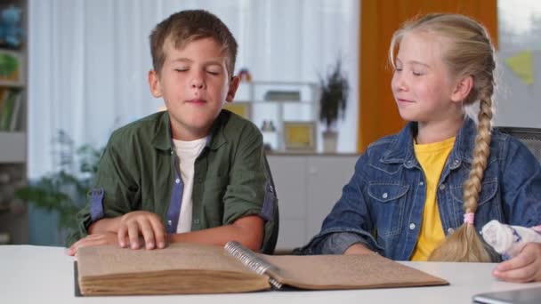 Niño lee libro usando sus manos en casa, niño ciego aprende mientras está sentado con su hermana con yeso fundido en el brazo — Vídeo de stock