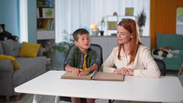 Слепой школьник дома, учительница учит слабовидящего мальчика читать книги Брайля с символами, сидя за столом — стоковое видео