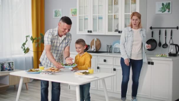 Familia amistosa junto con el niño varón conjunto de mesa para la cena conjunta en el fondo de la cocina — Vídeo de stock