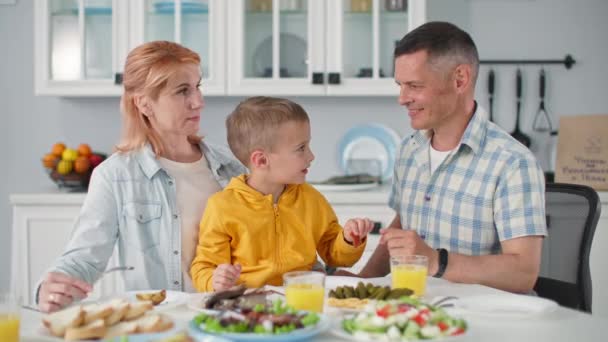 Gelukkige jeugd, vrolijke kleine jongen zit in mama armen en de hand voedt zijn glimlachende mannelijke ouder zitten aan tafel in de keuken tijdens het gezin — Stockvideo