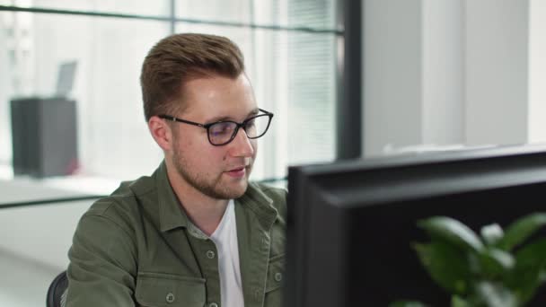 Портрет молодого чоловіка в окулярах, що розмовляють по телефону, працюючи за комп'ютером в офісі — стокове відео