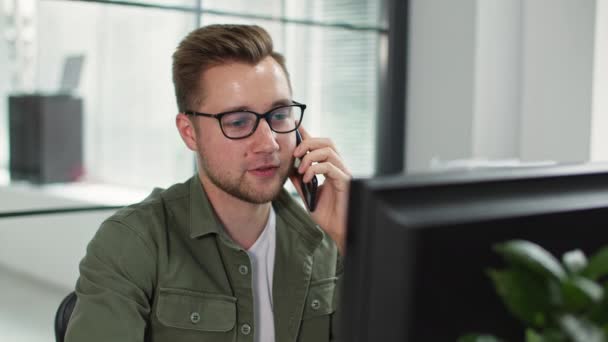 Portret młodego człowieka w okularach do wzroku rozmawiającego przez telefon komórkowy siedząc przy komputerze w biurze — Wideo stockowe