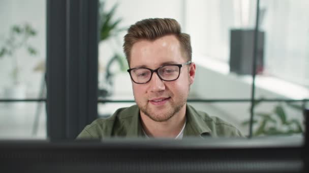 Молодой работник мужчина в очках для зрения работает за монитором, сидя за компьютером в офисе, крупным планом — стоковое видео