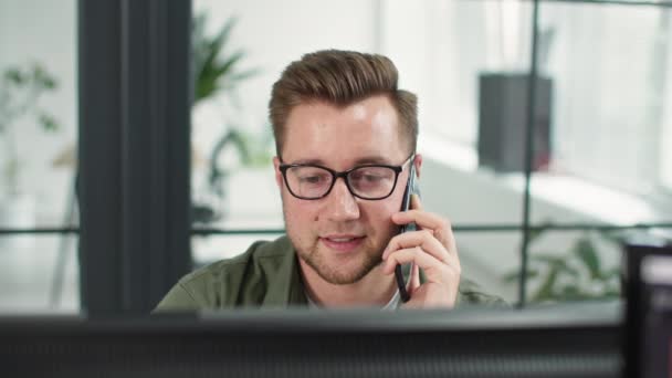 Усміхнений чоловік-працівник в окулярах розмовляє з клієнтом по телефону, працюючи за комп'ютером або отримуючи додатки, сидячи за столом в офісі — стокове відео