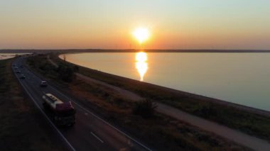 Hava aracı gün batımında Firth 'ün üzerinde uçuyor, deniz ve yol üzerinde hava manzarası yel değirmenleriyle büyüleyici manzaranın arka planında, gün batımında arabaların yanından geçiyor.