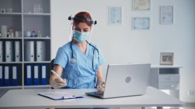 Yüzünde maske ve güneşlik olan doktorun uzaktan muayenesi hastane ofisinde otururken sağlıksız hastalara tavsiye veriyor.