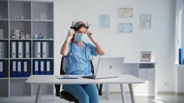 Precauciones, médico joven muestra en la cámara portátil cómo ponerse el casco protector y la máscara médica en la cara mientras está sentado en la oficina del hospital — Vídeos de Stock