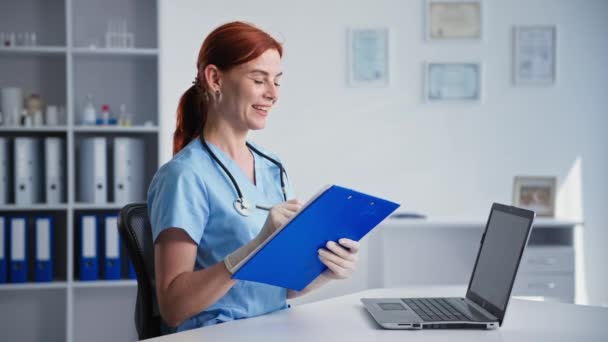 Портрет молодой женщины-врача в медицинской форме во время встречи с пациентом в режиме онлайн с помощью видеосвязи на ноутбуке, сидя в офисе клиники, улыбаясь и глядя — стоковое видео