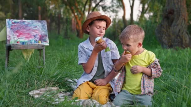 Happy joyful brothers are happy to eat a sweet cool summer dessert and lick their tongues ice cream while sitting on the lawn background of green trees — Stock Video