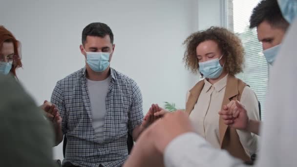 Religie, groep van jonge mannen en vrouwen in medische maskers houden hun vrienden handen en met gesloten ogen bidden tot God met hoop en geloof in hun hart terwijl ze zitten in een cirkel tijdens therapie sessie — Stockvideo