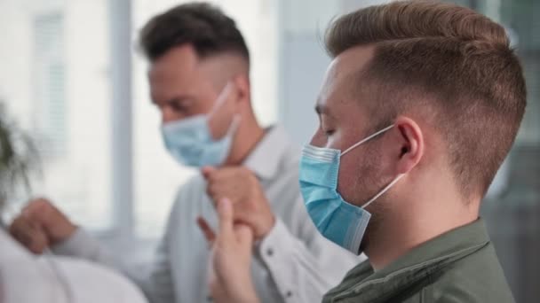 Jonge christelijke man draagt medisch masker neemt voorzorgsmaatregelen tijdens pandemie houden groep mensen door de handen tijdens collectief gebed tot God zitten in cirkel — Stockvideo