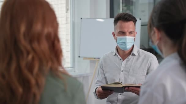 Młody człowiek przestrzega środków ostrożności i nosi maskę medyczną na twarzy, czytając Biblię z chrześcijańskim zespołem mężczyzn i kobiet siedzących w kółku. — Wideo stockowe
